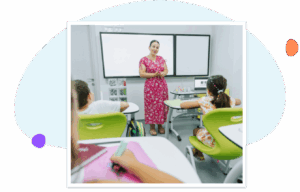 A smiling teacher in a pink floral dress stands at the front of a bright classroom, engaging with a small group of elementary students seated at green desks, with a whiteboard and laptop visible behind her.