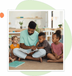 A teacher sits on the classroom floor reading a picture book to a diverse group of young children, who are attentively listening and gathered closely around him.