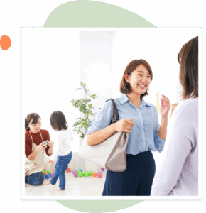 Alt text: Two women stand talking and smiling in a bright, welcoming classroom while young children play and interact with a caregiver in the background. The environment feels inviting and family-friendly, emphasizing communication and connection between adults and children.