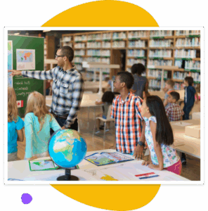 A teacher stands beside a group of elementary students in a library, pointing to a world map on a chalkboard while the children look on with interest. A globe and open atlases are on the table in the foreground, suggesting a lesson about countries and continents.